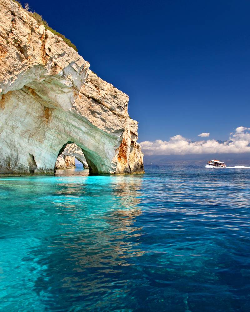 Crystal clear turquoise water laps at the base of a towering, sun-drenched cliff with an arched opening, while a small boat speeds across the deep blue sea in the distance under a bright sky.