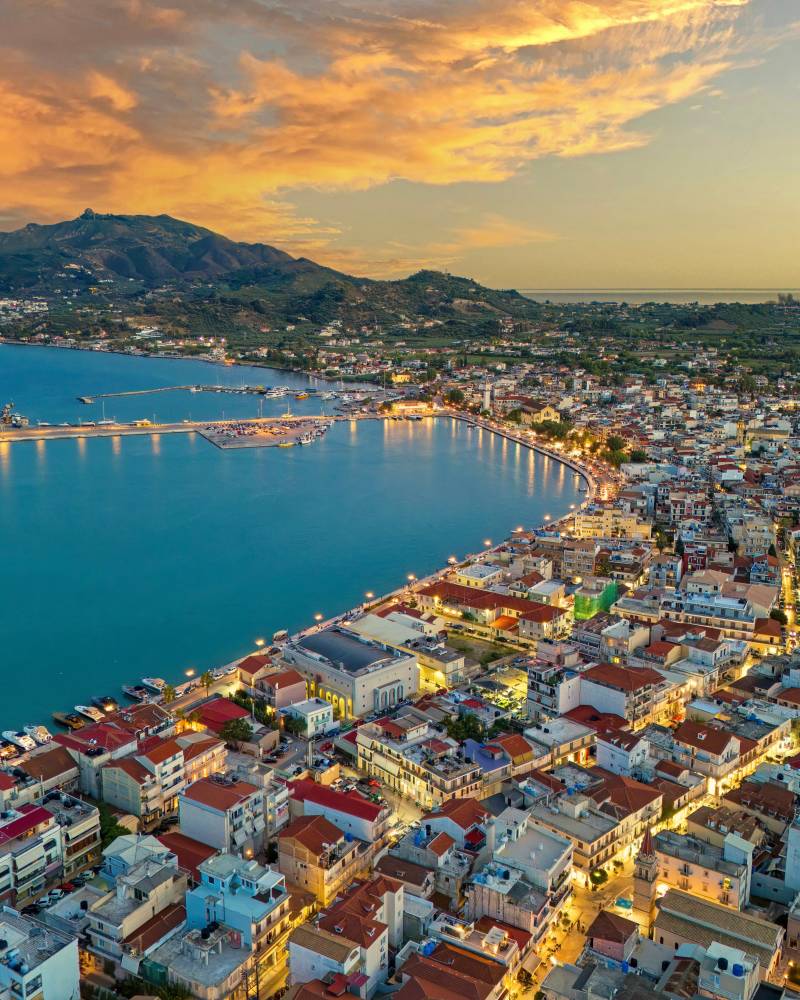 A coastal town with illuminated buildings in the foreground curves around a bay filled with boats, leading to a mountainous landscape under a colorful sunset sky.