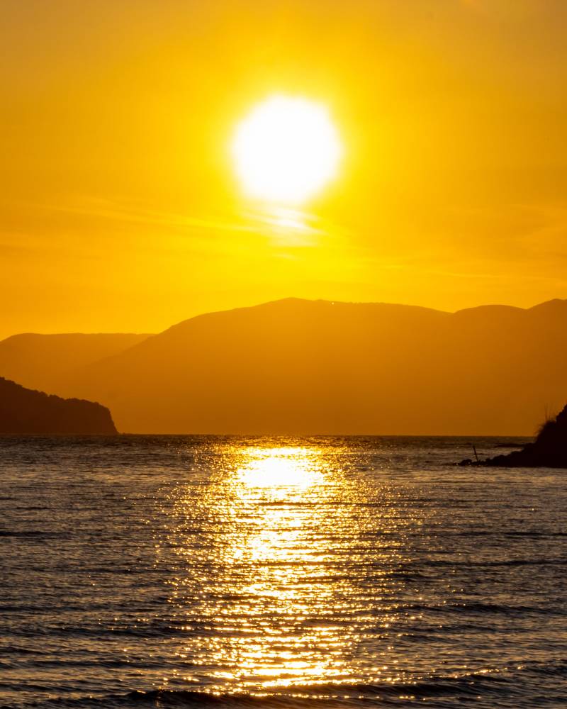 A brilliant sun sets over a calm sea, casting a golden reflection on the water, with silhouetted mountains in the distance.