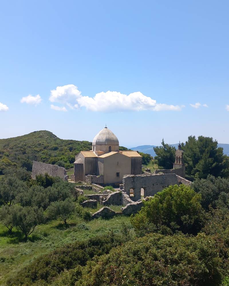 A domed stone building with surrounding ruins sits amidst lush green trees and rolling hills under a bright blue sky with scattered clouds.