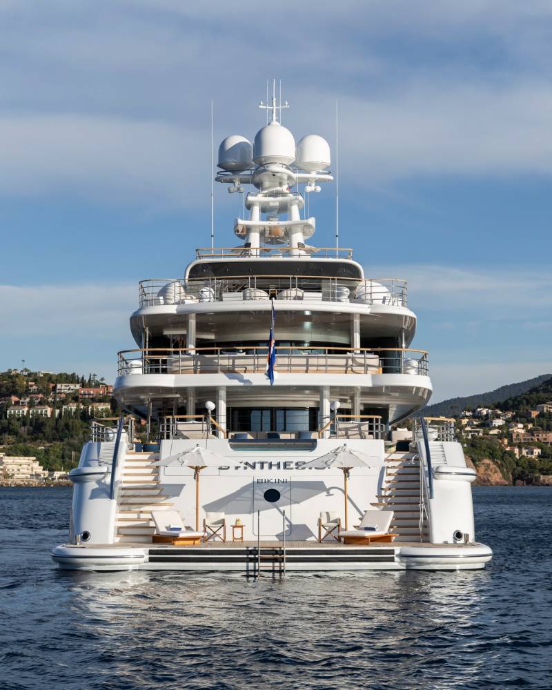 A large white yacht with multiple decks sits on calm blue water, with lush green hills dotted with buildings and a bridge visible in the background under a clear sky.
