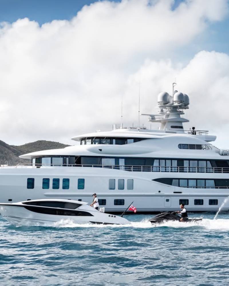 A large white yacht is anchored in the ocean, with two jet skis zipping around it, while lush green hills rise in the background under a cloudy sky.