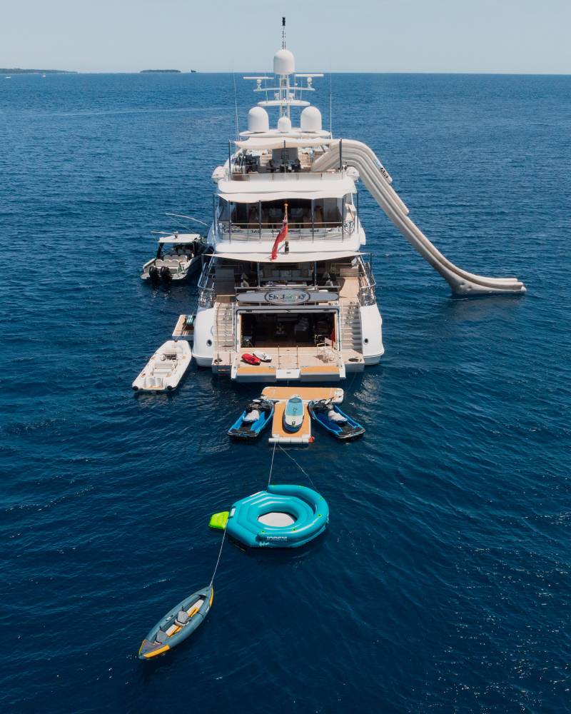 A large yacht with a waterslide and several smaller boats is anchored in the calm blue sea, with a rocky coastline visible in the distance.