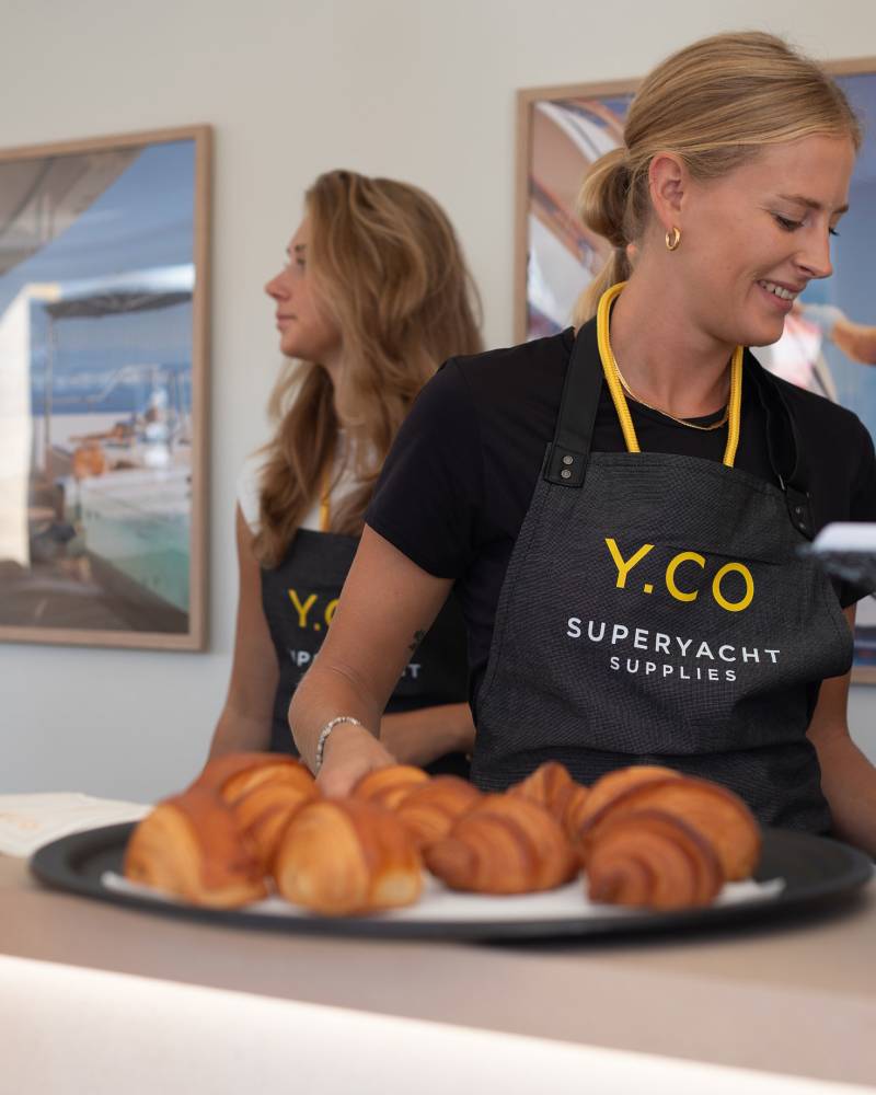 A smiling woman in a Y.CO apron serves coffee to a customer in the foreground, with another woman and framed artwork visible in the background.