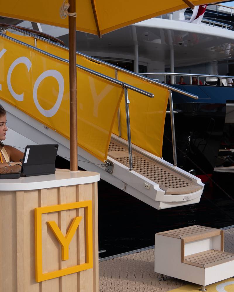 A woman works at a Y.CO. branded stand in the foreground, with a large yacht named "LADY BEATRICE" docked behind her.