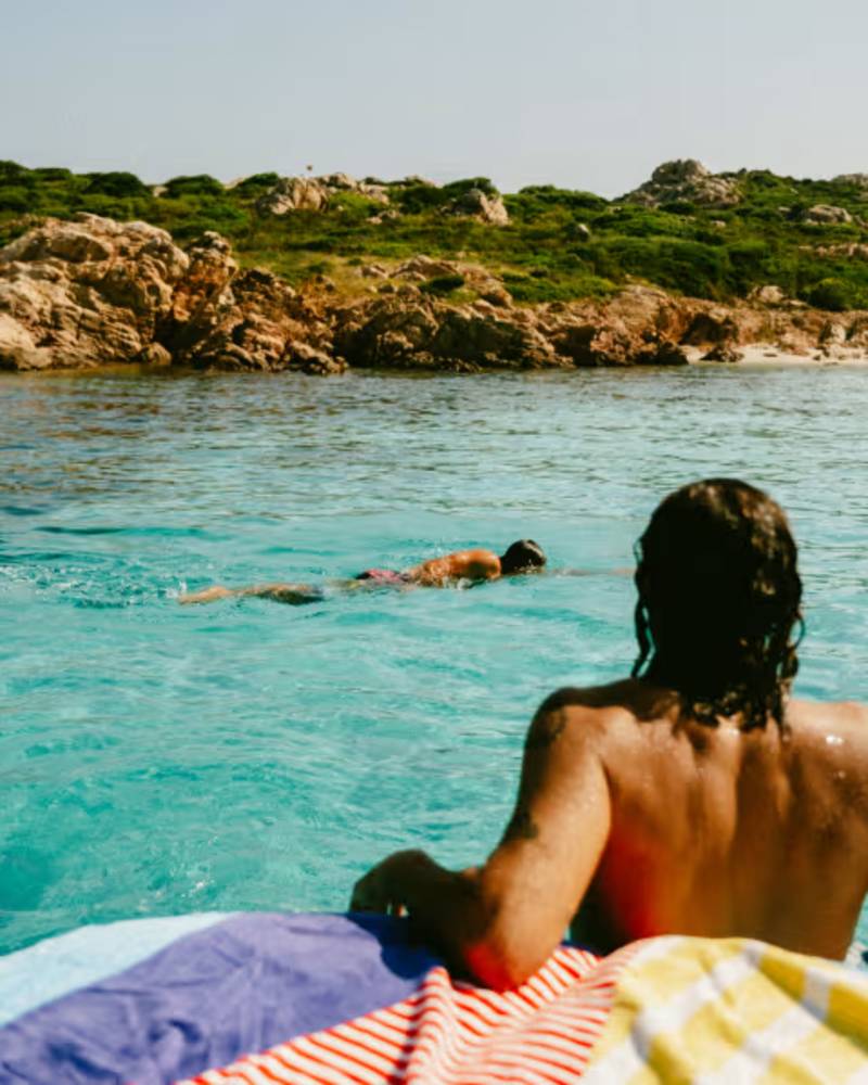 A person floats in clear turquoise water with a rocky, green hillside in the background, while another person rests on a colorful towel in the foreground.