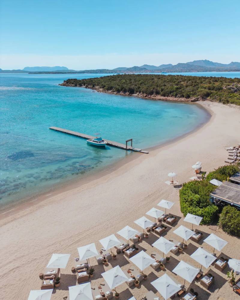 White umbrellas and lounge chairs dot a sandy beach in the foreground, leading to a calm turquoise bay with a wooden pier and a boat, all set against a backdrop of lush green hills under a clear blue sky.