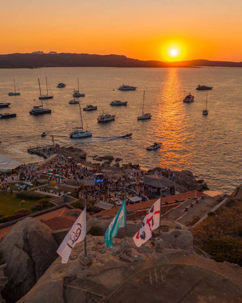 Flags wave in the foreground as a sunset illuminates a bay filled with boats and a lively gathering on the shore.