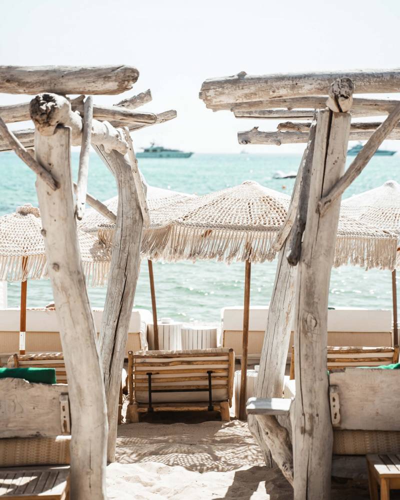 Weathered wooden structures frame a serene beach scene with lounge chairs and umbrellas in the foreground, leading to a calm ocean with yachts in the background.