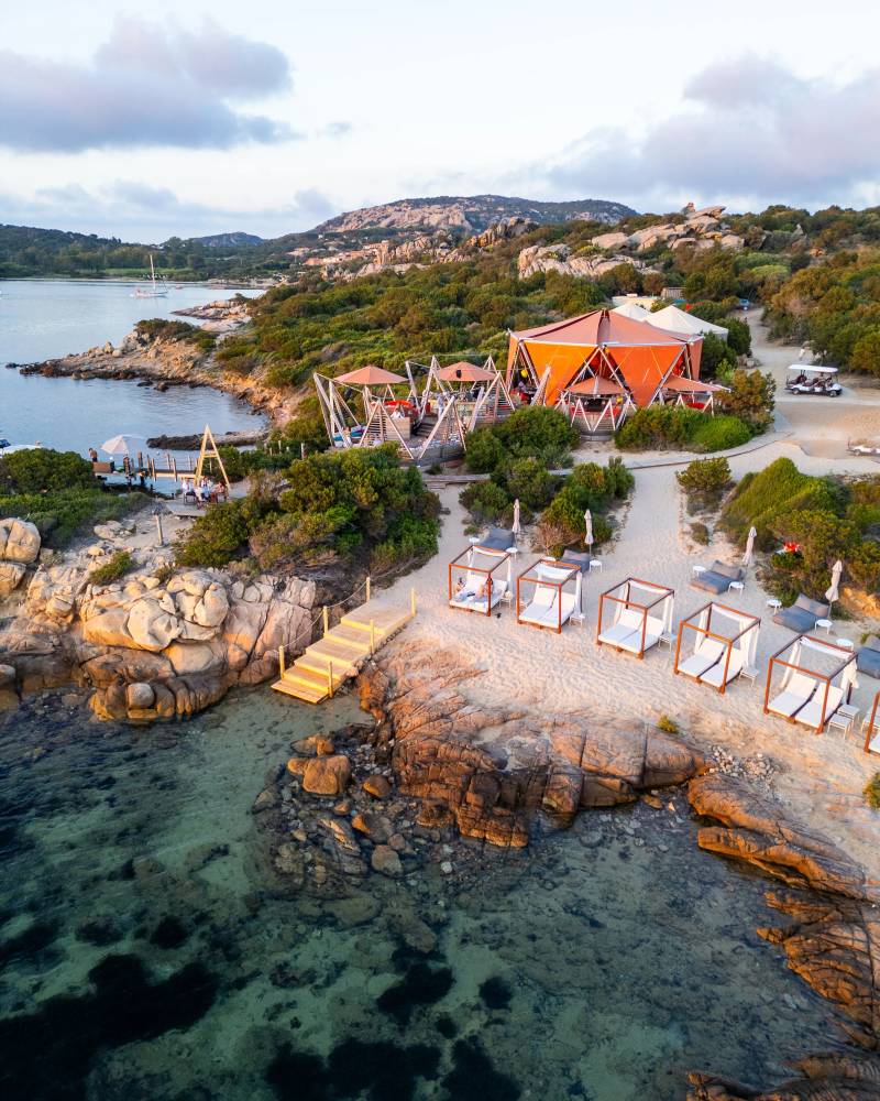 In the foreground, clear turquoise water laps against a rocky shore dotted with white cabanas, while the background features a vibrant orange building nestled amongst lush green foliage and rolling hills under a cloudy sky.