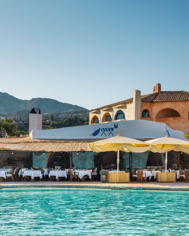 A bright blue swimming pool is in the foreground, with a restaurant featuring white-clothed tables and yellow umbrellas in the midground, and a building with arched windows and hills in the background.