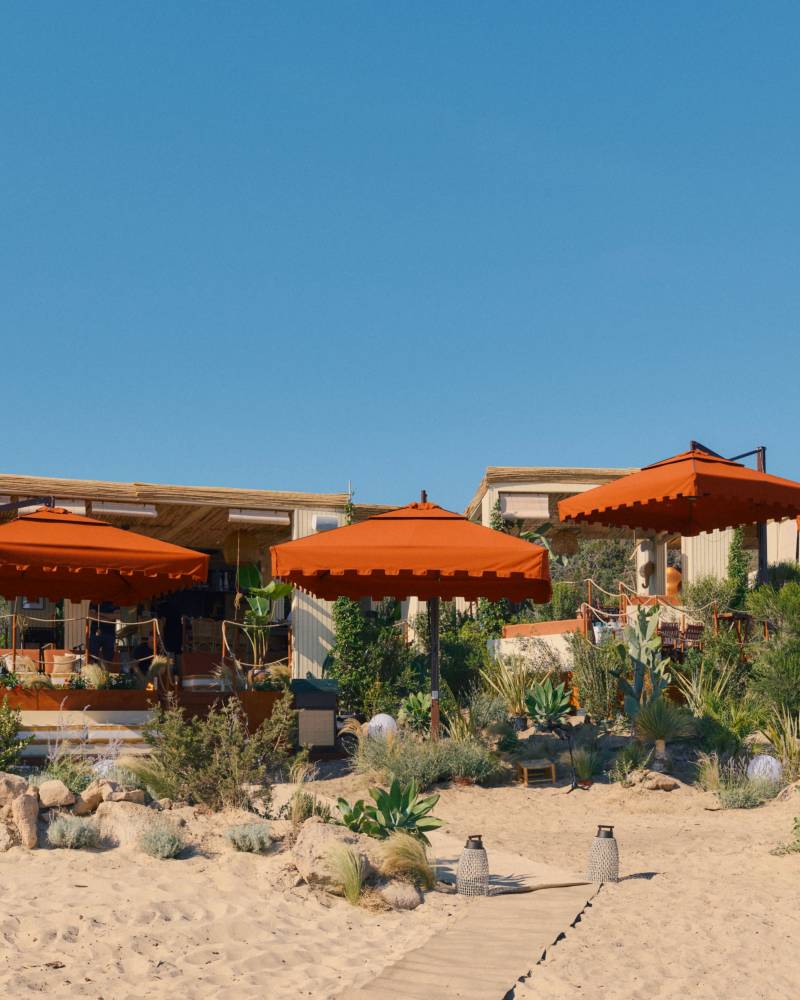 A sandy beach with a wooden path leads to a beachfront cafe with orange umbrellas, set against a clear blue sky.