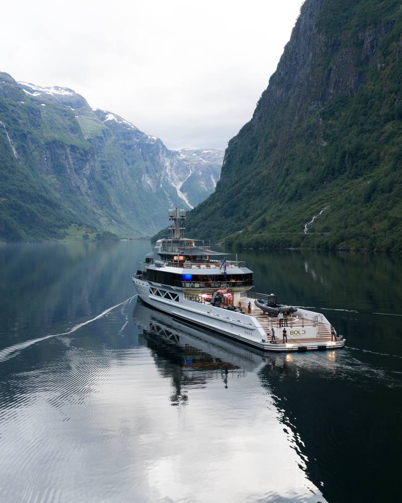 A luxurious yacht cruises through a serene, reflective fjord, flanked by towering, verdant mountains.
