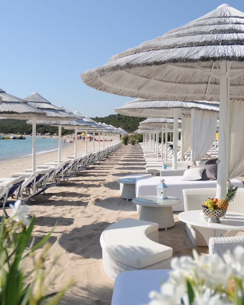 White lounge chairs and umbrellas line a sandy beach with calm blue water and distant green hills under a clear sky.