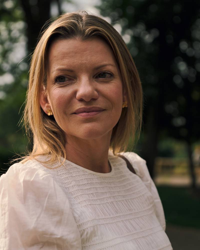 A blonde woman in a white top smiles softly in the foreground, with a blurred background of trees and foliage.