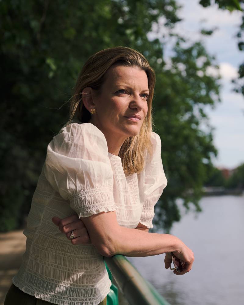A woman in a white blouse leans on a railing by a path, looking out at a calm river with trees in the background.