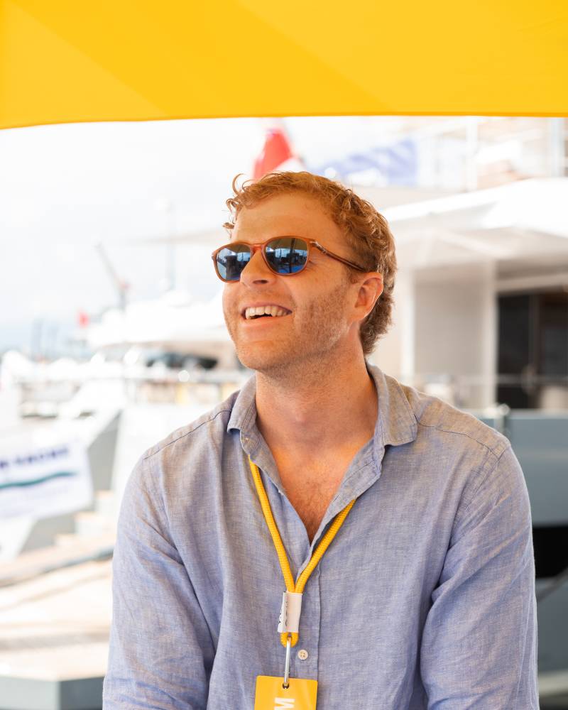 A man with red hair and sunglasses smiles while wearing a blue shirt and a yellow "TEAM" lanyard, with yachts visible in the blurred background under a yellow awning.