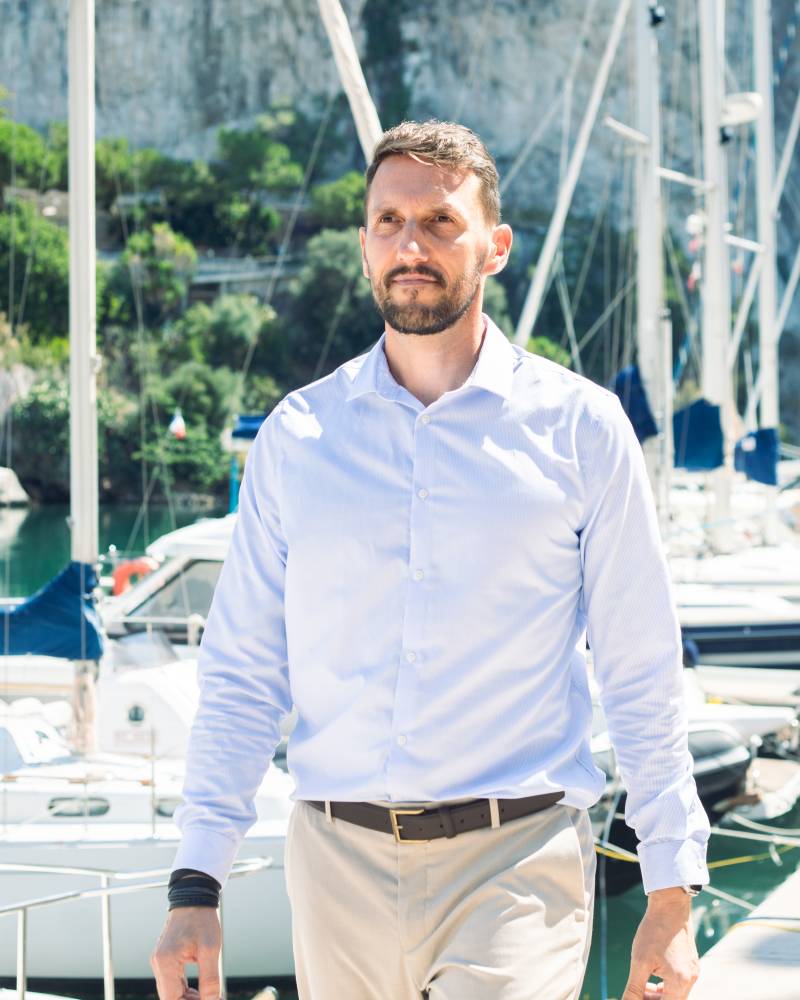 A man in a light blue shirt and tan pants walks along a dock with sailboats and a rocky cliff in the background.
