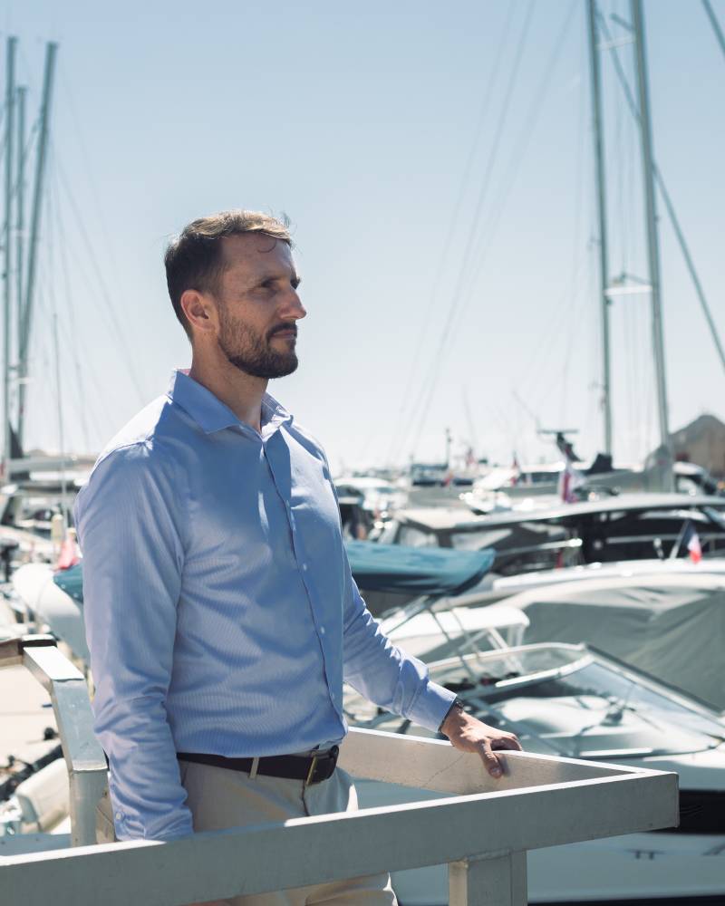 A man in a blue shirt stands on a dock, looking out at a harbor filled with sailboats and yachts under a clear sky.