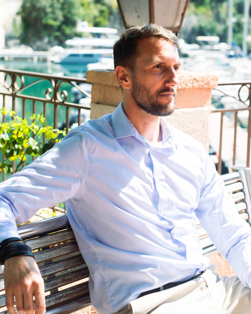 A man in a light blue shirt and beige pants sits on a bench in the foreground, with a harbor full of boats and lush greenery in the background.