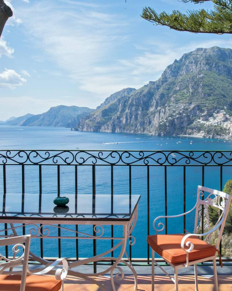 A table and chairs on a balcony overlook a sparkling blue sea and a picturesque coastal town nestled among green mountains.