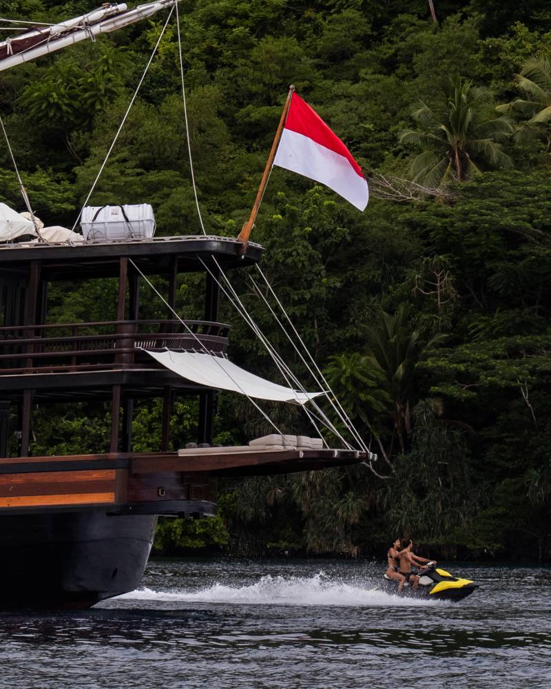 A jet ski speeds past a large wooden boat flying an Indonesian flag, with a lush green jungle in the background.
