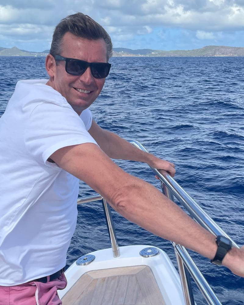 A man in a white t-shirt and pink shorts smiles while leaning on a railing on a boat, with a blue ocean and distant islands in the background.