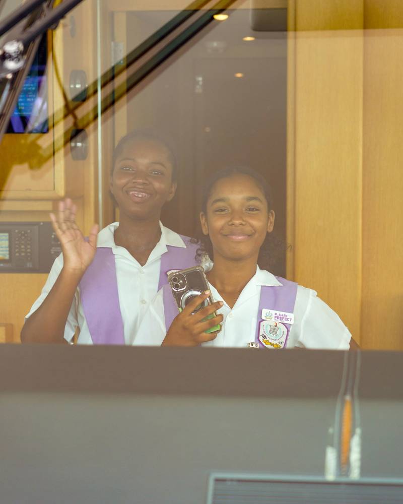 Two smiling girls in school uniforms wave from behind a glass partition, with a framed picture and wooden paneling visible in the background.