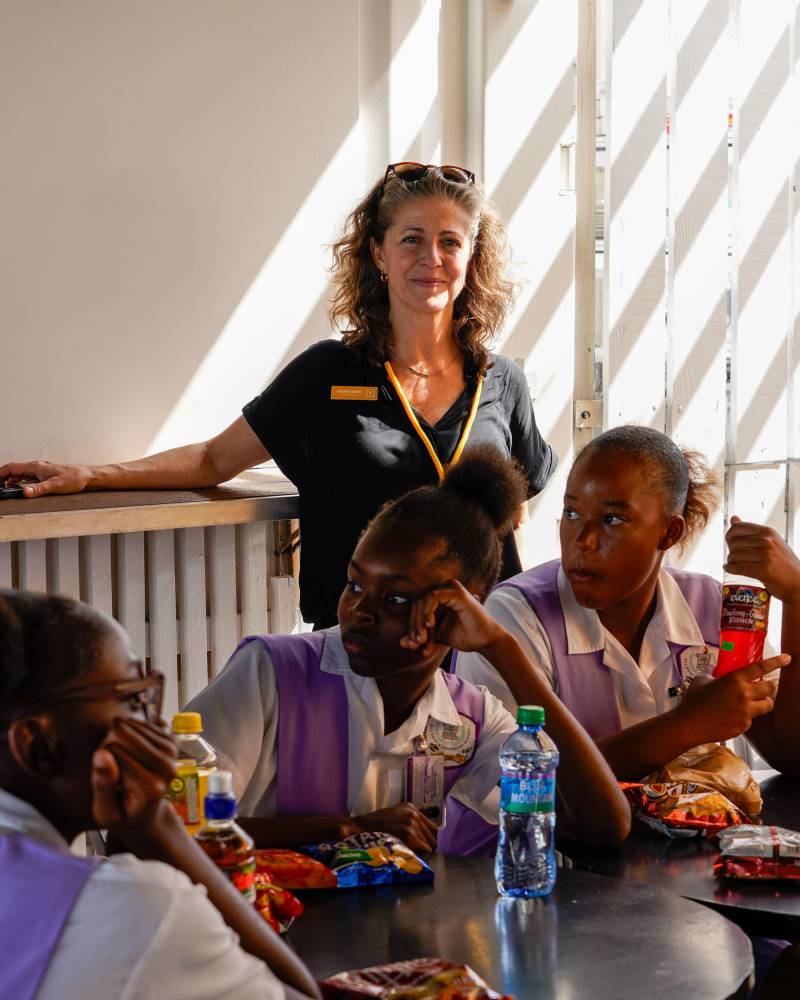 A woman stands behind a group of young people seated at a table with drinks and snacks, with a slatted wall in the background.
