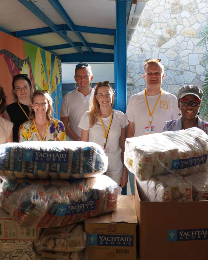 A group of volunteers stands behind stacks of donated food items in front of a colorful mural and palm trees.