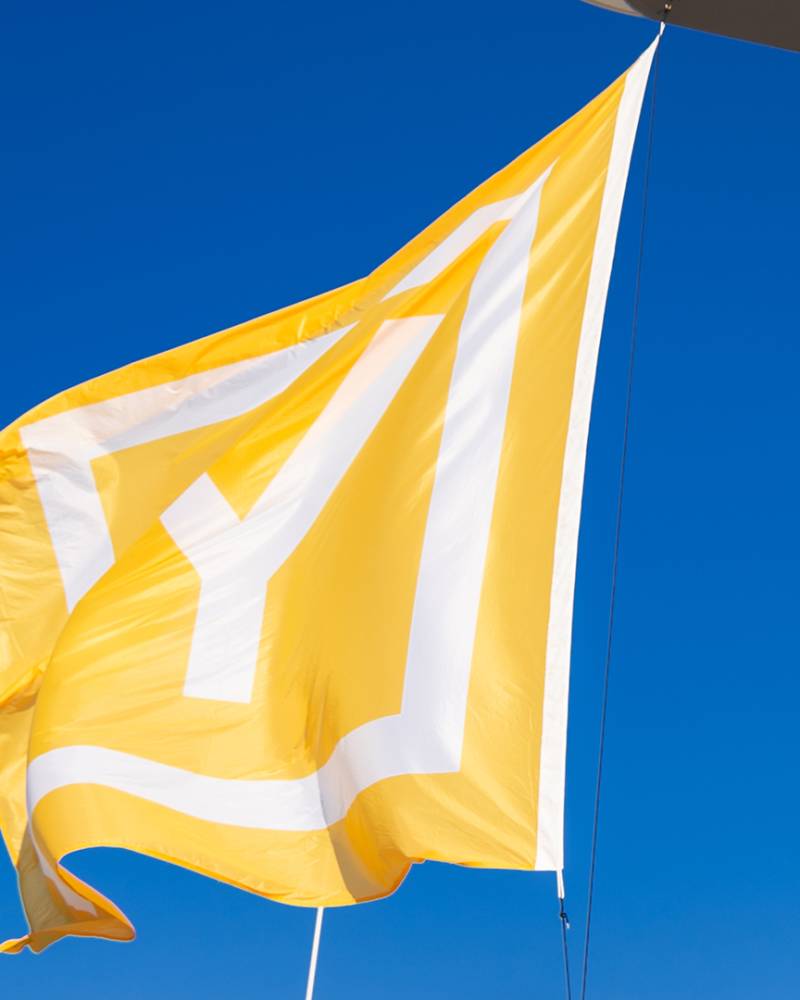 A yellow flag with white markings flutters against a clear blue sky, with the edge of a boat visible in the foreground.