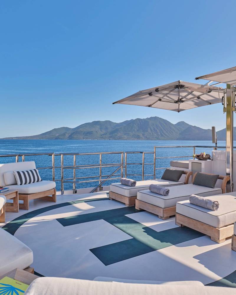 Sun loungers and seating areas on a yacht's deck overlook a calm sea and distant, mountainous coastline under a clear blue sky.