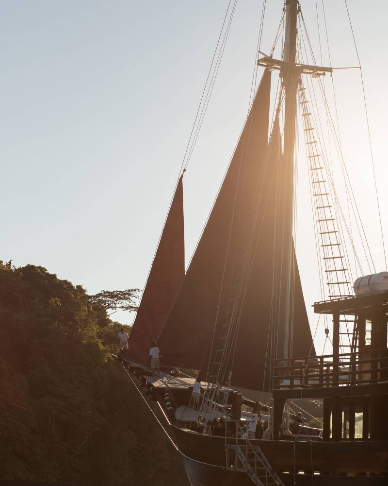 A large wooden sailboat with dark red sails is docked near a lush green island under a bright, hazy sky.