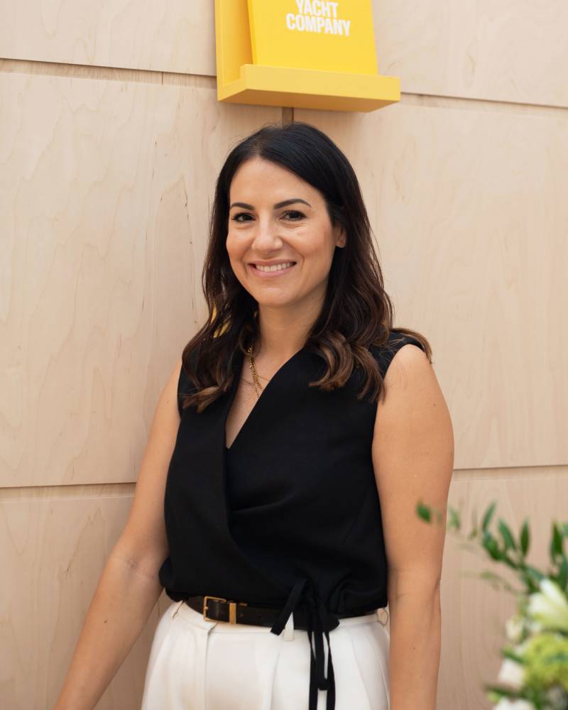A smiling woman in a black top and white pants stands in front of a light wood paneled wall with a yellow sign above her.