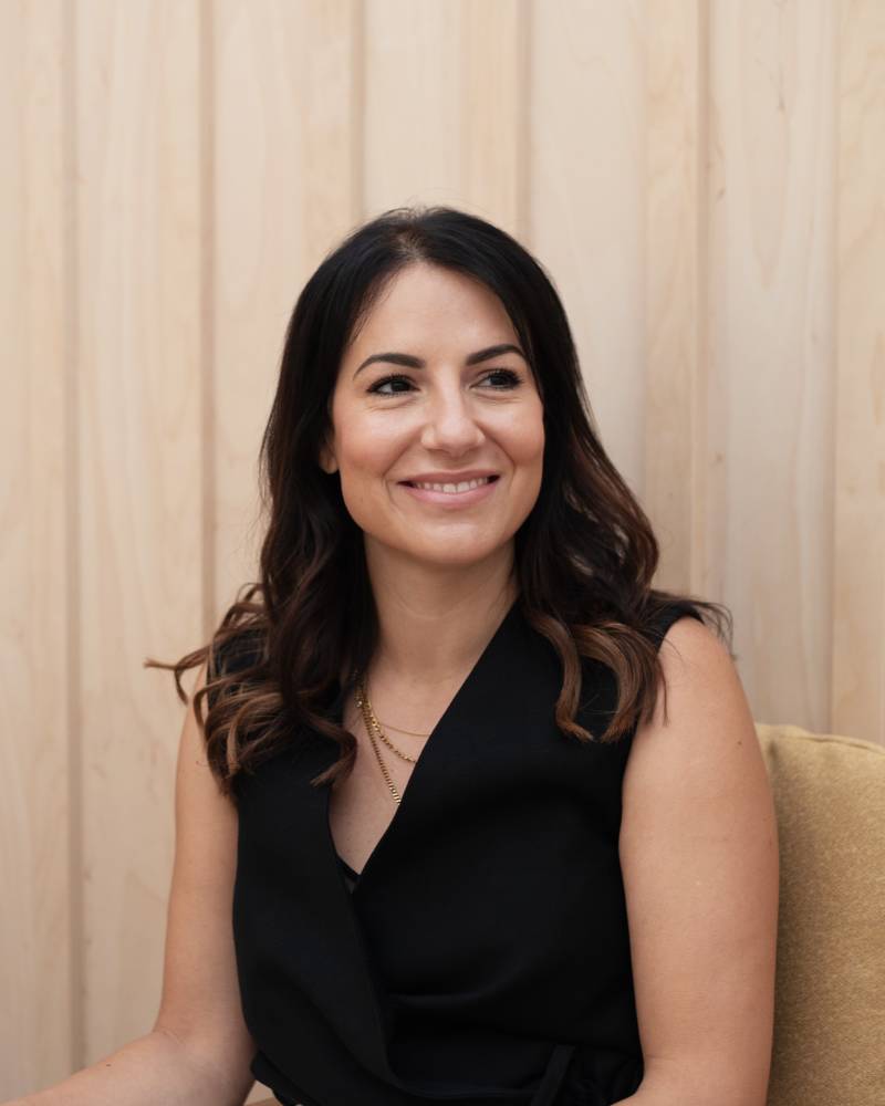 A woman with dark hair and a black top smiles while sitting in a chair against a light wood-paneled background.