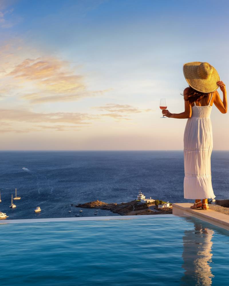 A woman in a white dress and straw hat stands by an infinity pool, holding a glass of wine, with a picturesque bay dotted with sailboats in the background under a sunset sky.