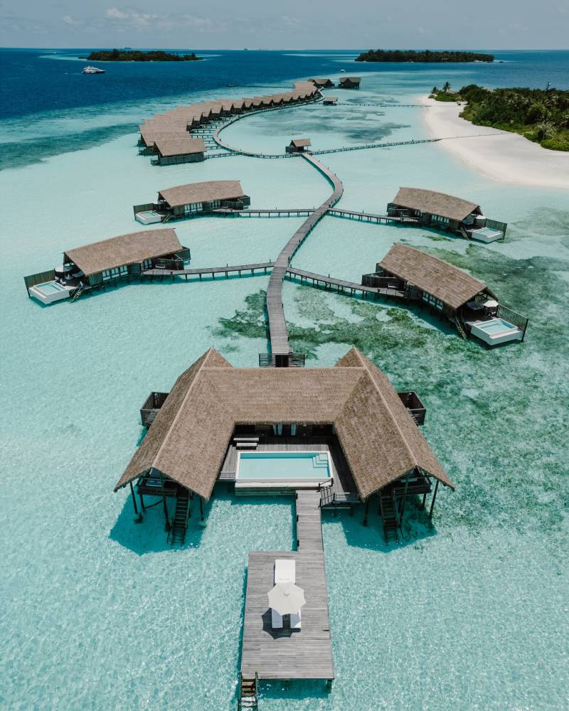 Overwater bungalows with private pools and decks extend from a central resort building into the turquoise ocean, with a white sand beach and distant islands in the background.