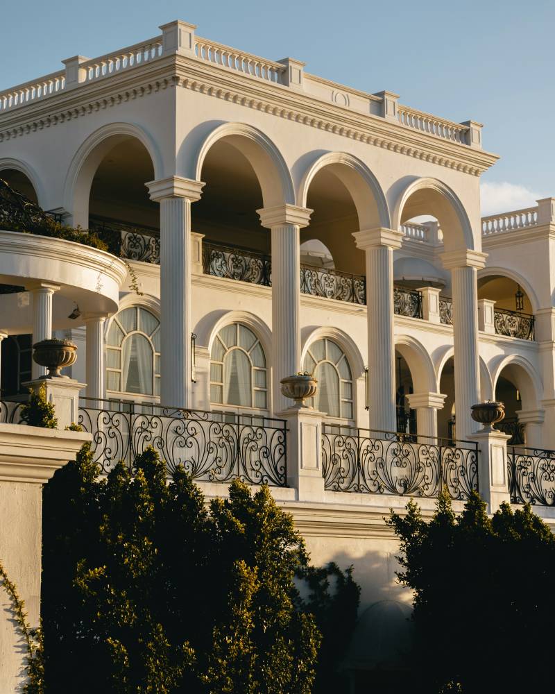 Lush green trees in the foreground frame a grand white building with arched windows and a decorative balustrade under a clear sky.