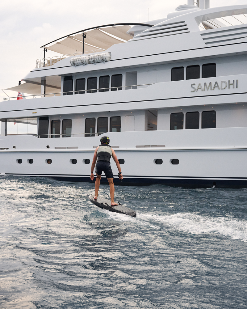 A person on a jetboard rides the waves in front of a large white yacht named Samadhi.