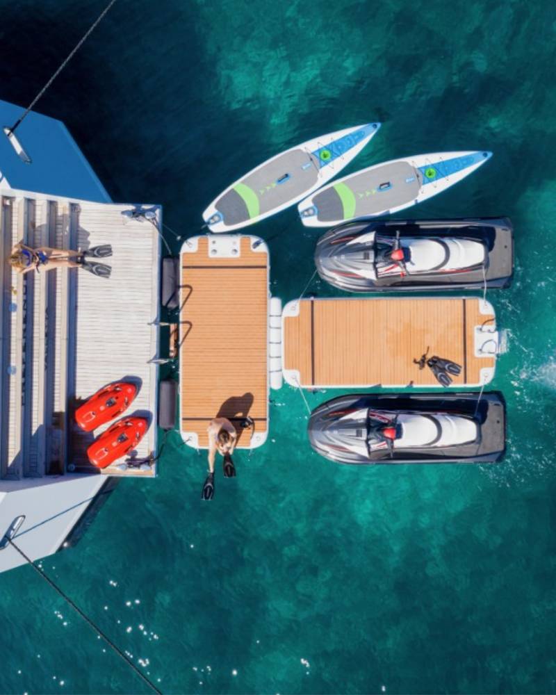 People relax on a yacht's deck and swim in the clear blue ocean alongside jet skis and paddleboards.
