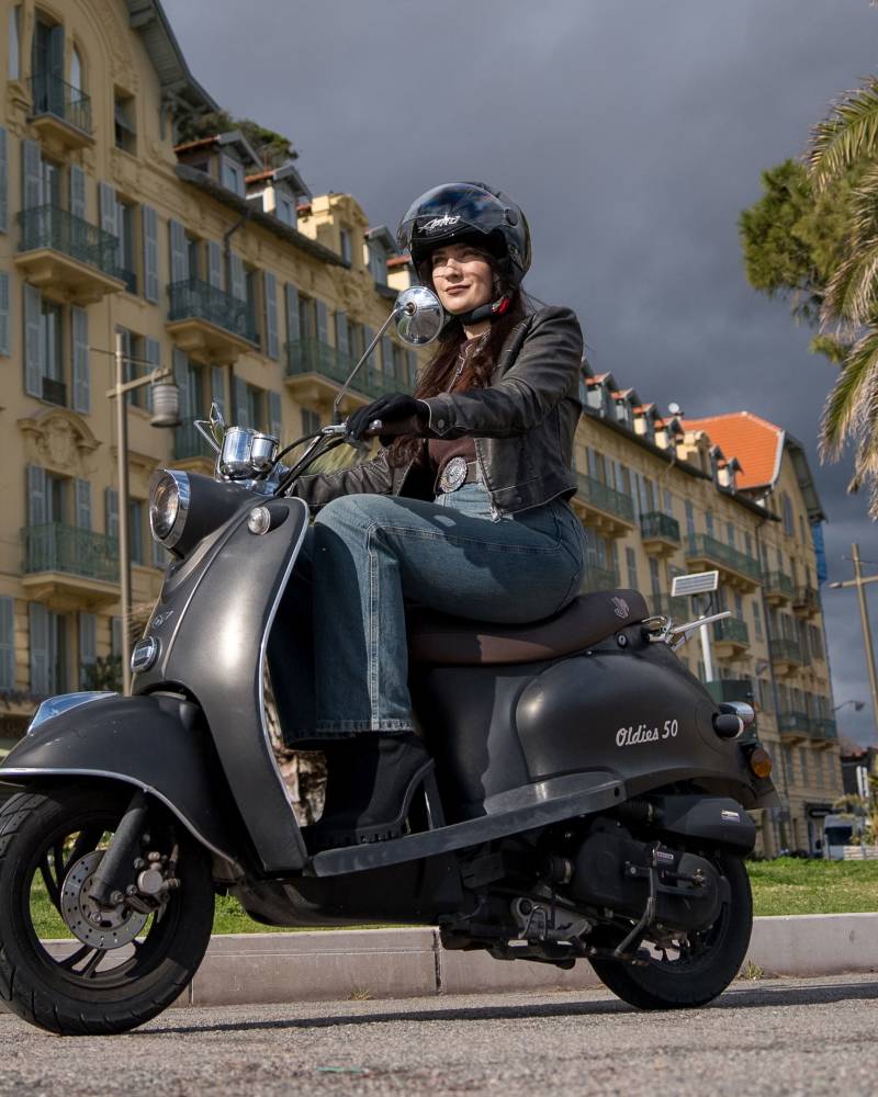 A woman in a helmet and leather jacket rides a dark gray scooter on a street with palm trees and ornate buildings in the background.