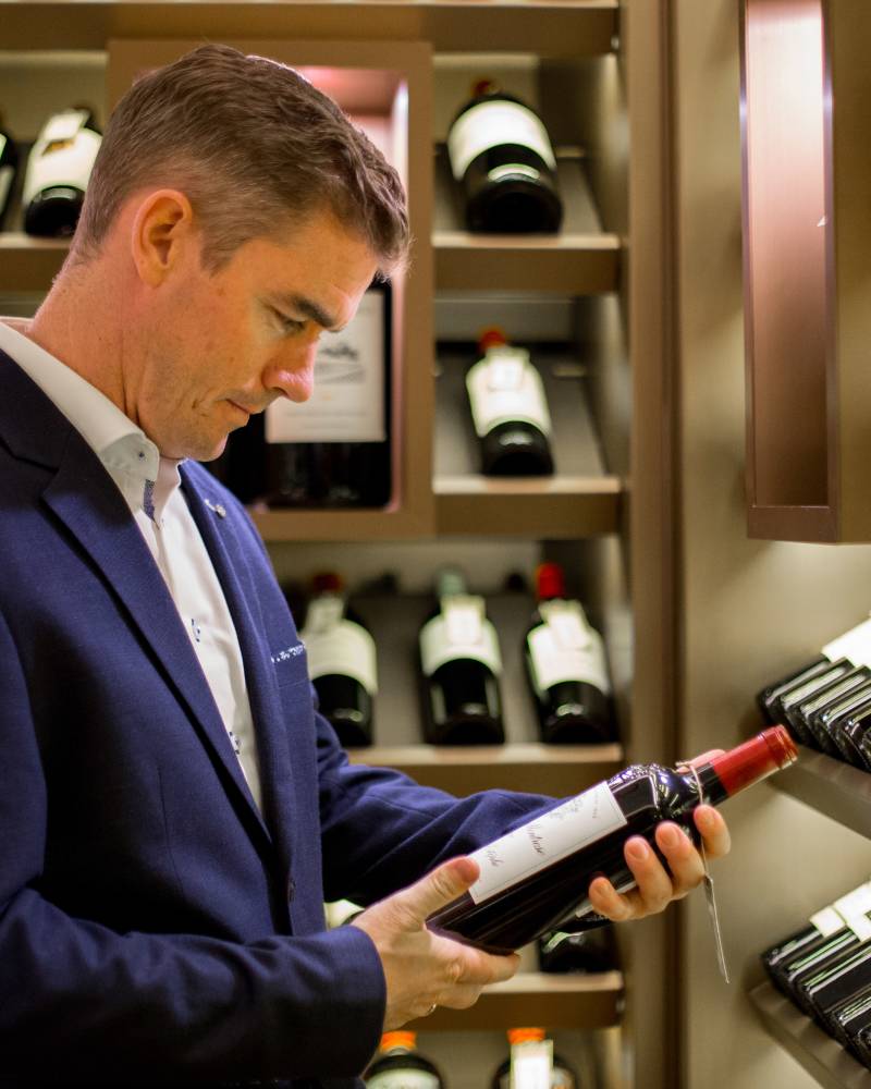A man in a suit examines a bottle of wine in the foreground, with shelves full of wine bottles lining the background.