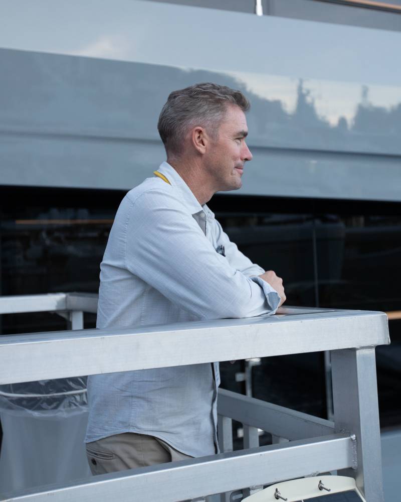 A man in a light blue shirt leans on a railing in the foreground, with a large gray yacht in the background.