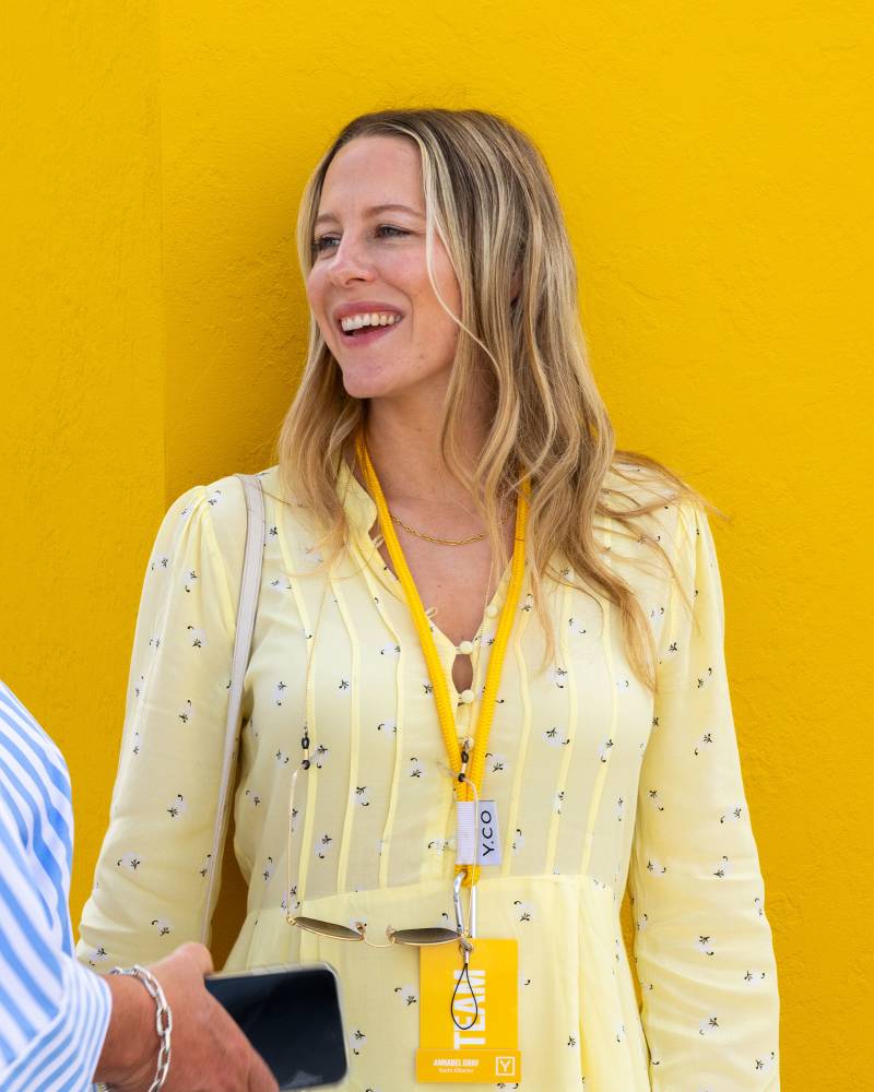 A smiling woman in a yellow dress with a lanyard stands against a bright yellow wall, with a person holding a phone in the foreground.