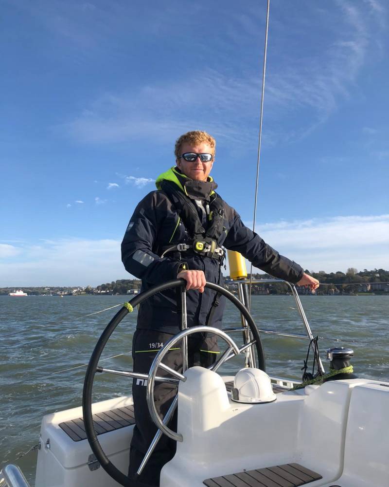 A young man in a life vest stands at the helm of a sailboat, with a clear blue sky and distant coastline in the background.