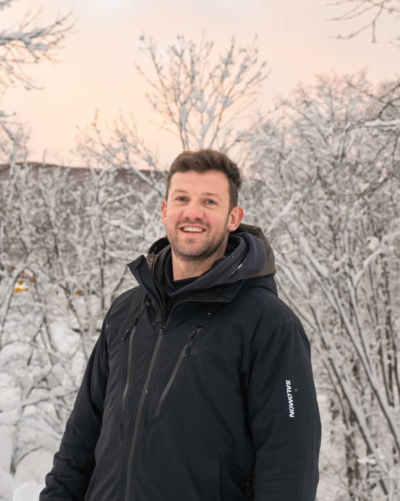 A smiling man in a black ski jacket stands in the foreground with snow-covered trees and a pale sky in the background.