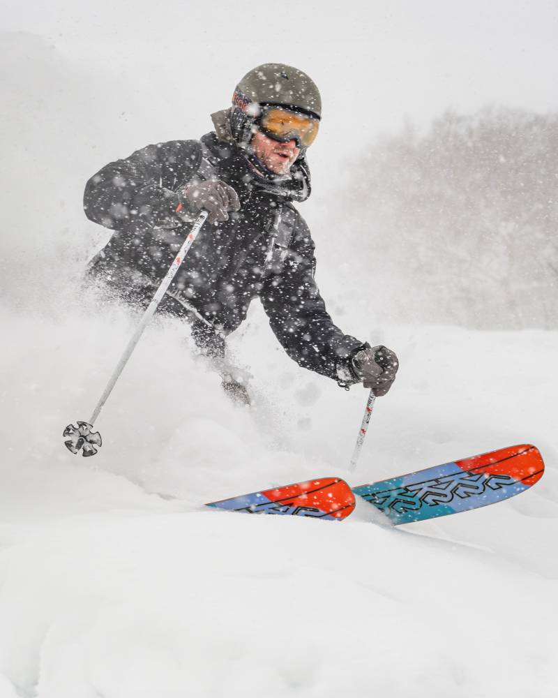 A skier carves through deep powder snow, with a blurred, snowy mountain landscape in the background.
