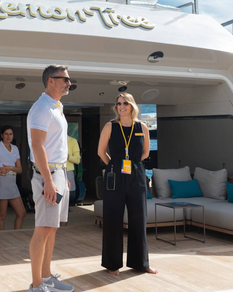 A man and a woman stand on the deck of a yacht, with a lounge area and the ocean visible in the background.