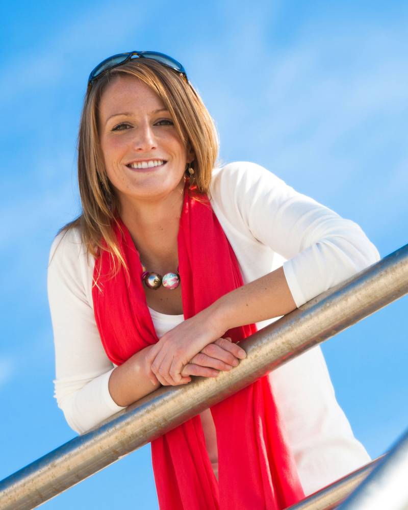 A smiling woman in a white top and red scarf leans on a metal railing against a bright blue sky.