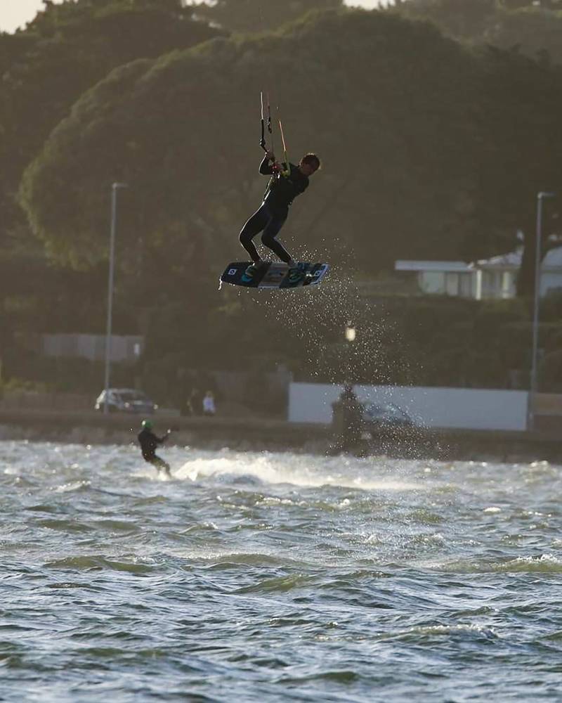 A kitesurfer soars through the air above choppy water, with a tree-lined street and buildings in the hazy background.